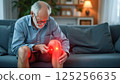 Elderly man using infrared device to relieve pain, inflammation on knee in cozy living room 125256635