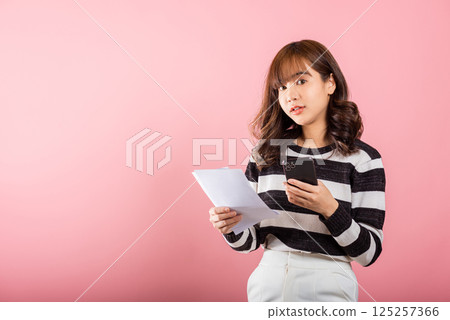 Happy Asian young woman holding mobile smart phone and family budget cost bills document invoice online for payment, Convenience online internet banking. female hold work papernote 125257366