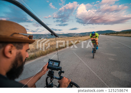A videographer recording a triathlete riding his bike preparing for an upcoming marathon.Athlete's physical endurance and the dedication required to succeed in the sport. A videographer recording a triathlete riding his bike preparing for an upcoming marathon.Athlete's physical endurance and the dedication required to succeed in the sport. 125258674