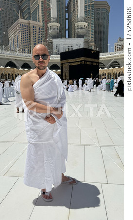 A bald man poses for a photo in front of the Kaaba at noon after performing Umrah at Masjid al-Haram, Mecca. A sacred Islamic pilgrimage in the heart of the holy mosque under the bright sunlight. A bald man poses for a photo in front of the Kaaba at noon after performing Umrah at Masjid al-Haram, Mecca. A sacred Islamic pilgrimage in the heart of the holy mosque under the bright sunlight. 125258688