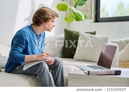 Teenager guy student studying sitting at home on sofa using laptop textbook notebook Teenager guy student studying sitting at home on sofa using laptop textbook notebook 125259597