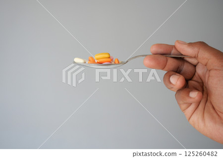 Hand holding a spoon with various colorful pills in plain setting 125260482