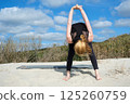 Woman practicing yoga outdoors on sandy surface, performing forward bend with clasped hands behind back, embracing balance, flexibility, and mindfulness under bright sky and nature 125260759