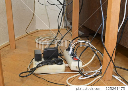 Tangled cables on the parquet floor. Overloaded electrical boards on the wooden floor. Electrical cables in disorder. 125261475