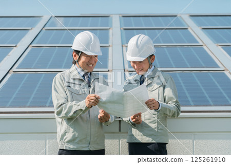 An engineer in work clothes from an electric power company or electrical contractor standing in front of a solar power generation/solar panel (clean energy) An engineer in work clothes from an electric power company or electrical contractor standing in front of a solar power generation/solar panel (clean energy) 125261910