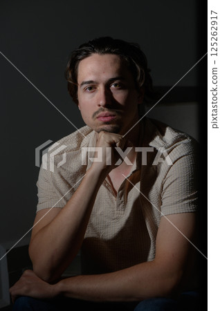 Reflective portrait of a young man in soft lighting, showcasing deep thoughts and intrigue in a cozy indoor space 125262917