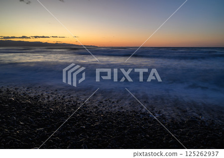 Glistening waves embrace stony shores as twilight descends over the horizon in a tranquil coastal paradise 125262937