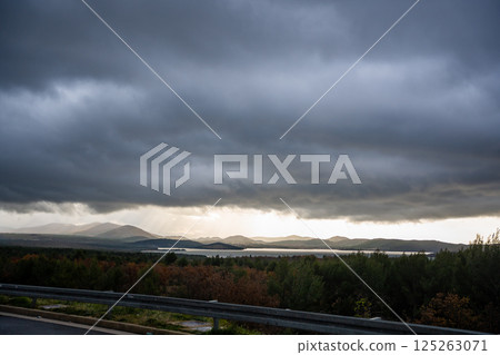 Scenic roadside view in Croatia with rolling hills, dense forests, and a shimmering lake under dramatic cloudy sky. Concept of road trip exploration, untouched nature, breathtaking travel landscapes 125263071