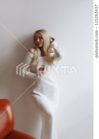 Elegant blonde in a white knitted dress stands near a white wall 125263437