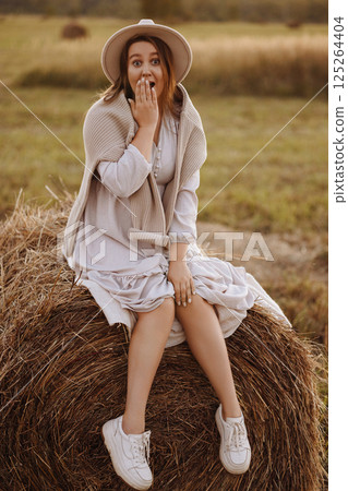 Young woman in beige dress sitting on haystack in village field Young woman in beige dress sitting on haystack in village field 125264404