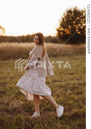 Front view of a young adult woman in a white dress in a field at sunset. 125264415