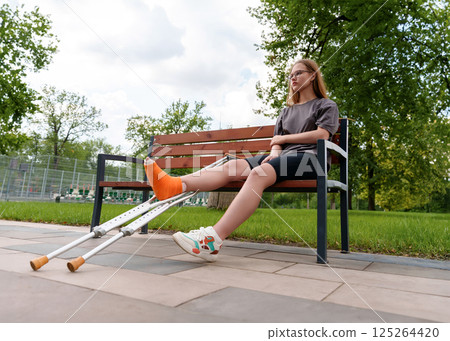 Pensive teenage girl, her leg in a cast, takes a seat on a park bench amidst the calm summer atmosphere, her crutches nearby 125264420