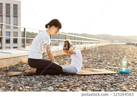 Mother helps young daughter perform abdominal exercises on rooftop during a sunny day, showcasing health and fitness. Mother helps young daughter perform abdominal exercises on rooftop during a sunny day, showcasing health and fitness. 125264563