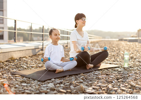 Mother and daughter sitting outdoors on rooftop doing exercises with dumbbells during sunrise. Both models smiling wearing casual sportswear promoting family fitness, outdoor activity Mother and daughter sitting outdoors on rooftop doing exercises with dumbbells during sunrise. Both models smiling wearing casual sportswear promoting family fitness, outdoor activity 125264568