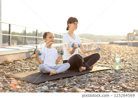 Caucasian mother and young daughter exercising with dumbbells, promoting fitness, sunny morning outdoors. 125264569