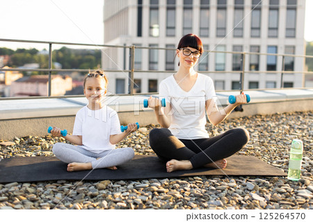 Mother and young daughter sitting on rooftop together exercising holding small dumbbells. Bright casual setting promoting healthy lifestyle and family relationship. Mother and young daughter sitting on rooftop together exercising holding small dumbbells. Bright casual setting promoting healthy lifestyle and family relationship. 125264570