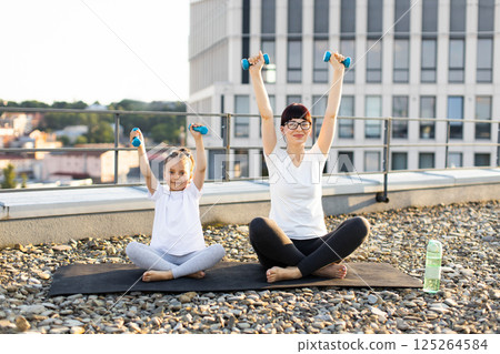 Mother and daughter sitting outdoors on rooftop practicing exercise with dumbbells, embracing fitness and bonding. Bright and sunny morning, both engaging in healthy lifestyle activities. 125264584
