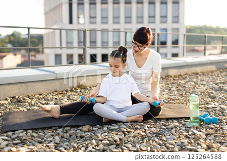 Mother sitting with daughter on rooftop, practicing fitness exercises using small dumbbells, enjoying bonding time. Child learning proper exercise techniques from parent. Mother sitting with daughter on rooftop, practicing fitness exercises using small dumbbells, enjoying bonding time. Child learning proper exercise techniques from parent. 125264588