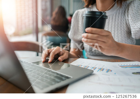 Professional woman multitasking with laptop and coffee, reviewing financial documents in a contemporary office environment. 125267536