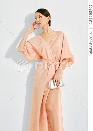 Elegant woman in a soft peach dress striking a pose with a clutch in a minimalist setting 125268791