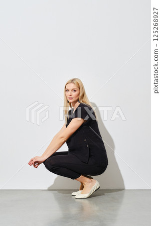 Woman squatting in profile next to a white wall 125268927