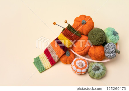 Green and orange knitting balls, orange pumpkins, striped scarf in a basket with knitting needles on beige background. Set for the hobby of knitting warm clothes or toys. Concept needlework 125269473