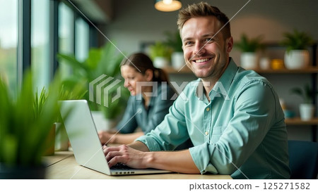 man smiling and looking at camera while working on laptop in modern office with green indoor plants and woman colleague, coworking, digital workspace, corporate communication 125271582
