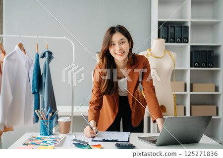 Successful fashion designer. Attractive young asian woman with smile while standing in workshop. Beautiful owner business woman working and holds tablet, laptop and smartphone in studio. Successful fashion designer. Attractive young asian woman with smile while standing in workshop. Beautiful owner business woman working and holds tablet, laptop and smartphone in studio. 125271836