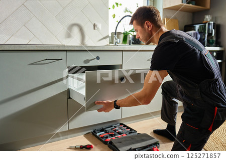 Man assembling kitchen drawer in modern cabinet using screwdriver. Handyman installing and adjusting drawer slide mechanism in contemporary kitchen. Furniture installation 125271857