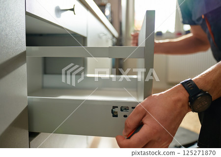 Man assembling kitchen drawer in modern cabinet using screwdriver. Handyman installing and adjusting drawer slide mechanism in contemporary kitchen. Furniture installation 125271870