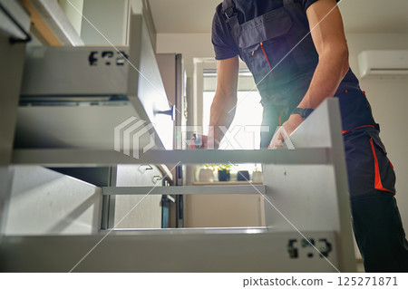 Man assembling kitchen drawer in modern cabinet using screwdriver. Handyman installing and adjusting drawer slide mechanism in contemporary kitchen. Furniture installation 125271871