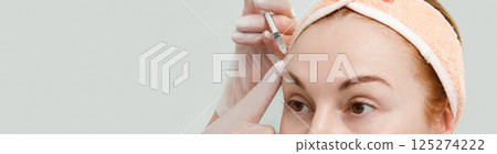 A close-up photograph of a middle-aged woman in which a doctor inserts a syringe into her forehead as part of a rejuvenation and wrinkle smoothing procedure. against the background of a white clinic 125274222