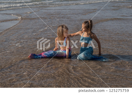 Little sister girls in mermaid costumes on the beach. Sunset on the shore of the blue sea, a child in a swimsuit. Childhood happiness, beach, vacation, sunscreen, warm weather, travel. High quality Little sister girls in mermaid costumes on the beach. Sunset on the shore of the blue sea, a child in a swimsuit. Childhood happiness, beach, vacation, sunscreen, warm weather, travel. High quality 125275759