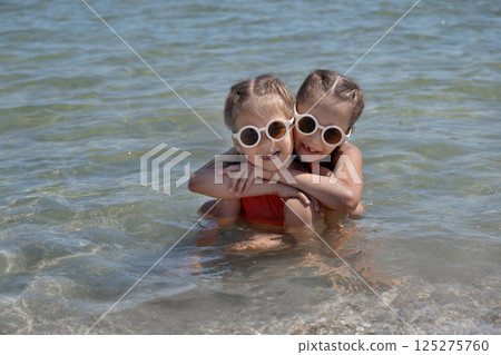 Girls in swimsuits, white sunglasses and hats on the beach. Sunset on the shore of the blue sea, a child in a swimsuit. Childhood happiness, beach, vacation, sunscreen, warm weather, travel. High 125275760