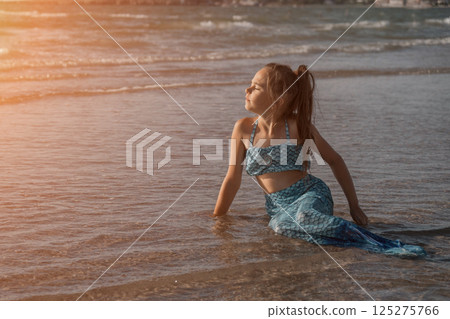 A girl in a mermaid costume on the beach. Sunset on the shore of the blue sea. Childhood happiness, beach, vacation, sunscreen, warm weather, travel. High quality photo 125275766