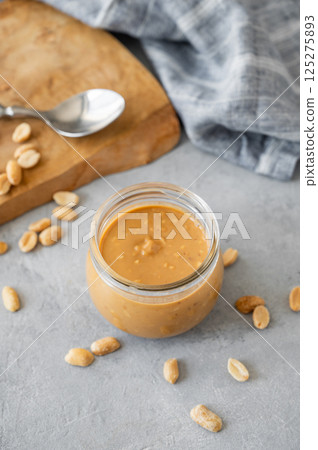 Glass jar filled with creamy peanut butter on grey background with scattered roasted nuts 125275893