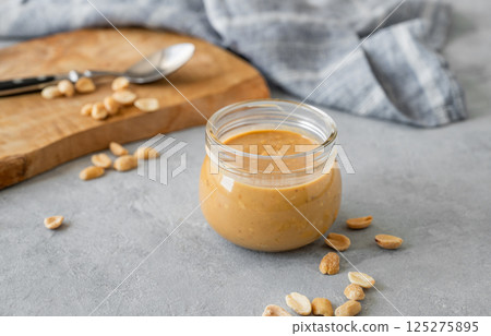 Glass jar filled with creamy peanut butter on grey background with scattered roasted nuts 125275895