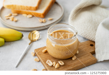 Glass jar filled with creamy peanut butter on grey background with bananas and fresh bread. 125275900