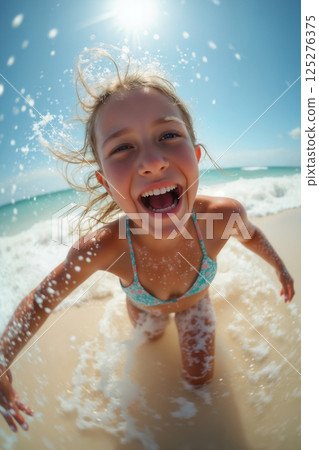 portrait of active child girl wearing bikini swimsuit, playing with water splashes and posing on resort beach with view of sea and bright sky 125276375