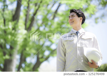 Close-up of the upper body of a man wearing work clothes and holding a helmet outdoors in fresh greenery under a blue sky. Image of construction management, work site, etc. 125276499