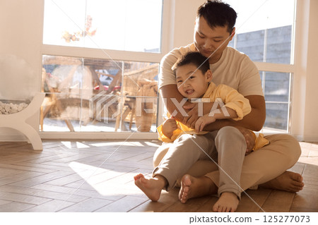 A young happy Asian man hugs his little son. A Korean dad is playing with his child on the living room floor. A young happy Asian man hugs his little son. A Korean dad is playing with his child on the living room floor. 125277073