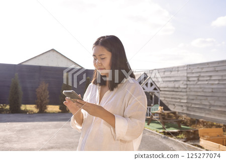 An attractive Asian holds her phone and types a message. A charming Korean girl in white clothes stands with a gadget in the backyard of the house. An attractive Asian holds her phone and types a message. A charming Korean girl in white clothes stands with a gadget in the backyard of the house. 125277074