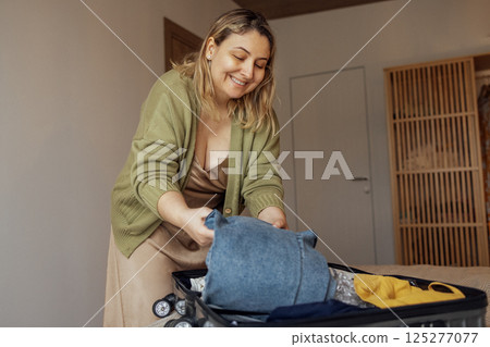 A young female blonde in a beige dress and a green jacket smiles and puts clothes in a suitcase in the bedroom in the house. A young female blonde in a beige dress and a green jacket smiles and puts clothes in a suitcase in the bedroom in the house. 125277077