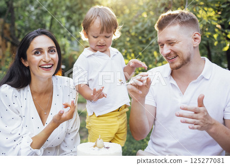 Little cute kid and his parents are tasting festive cake outdoors in garden. Happy family having fun. 125277081