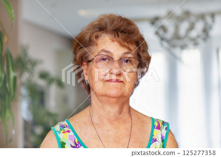 Confident elderly woman with short curly hair wearing glasses and dress, standing indoors at nursing home, looking calmly at camera. Wisdom, grace and positive aging 125277358