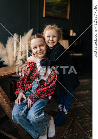 Vertical portrait of happy younger sister warmly hugging older sibling from behind, displaying intimate family connection with genuine emotional closeness inside comfortable home interior. 125277366