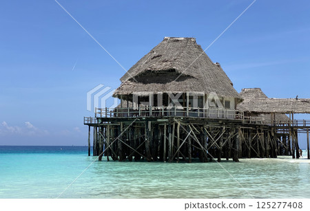 Beautiful tropical overwater bungalow with a thatched roof on stilts above clear turquoise waters. 125277408
