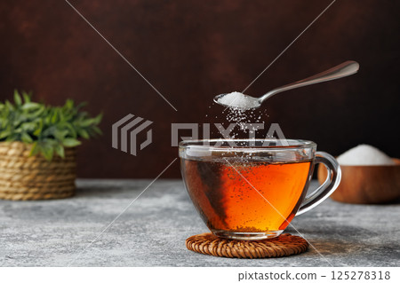 White sugar falling from teaspoon into glass cup of hot tea placed on coaster against dark brown background 125278318