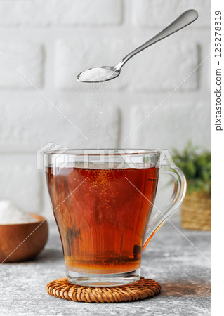White sugar falling from teaspoon into glass cup of hot tea placed on wicker coaster against brick wall as background 125278319