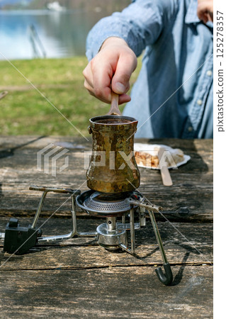 Person brewing traditional Turkish coffee in copper cezve on portable camping stove. Outdoor picnic near lake wooden table surface. Closeup hand stirring coffee. 125278357